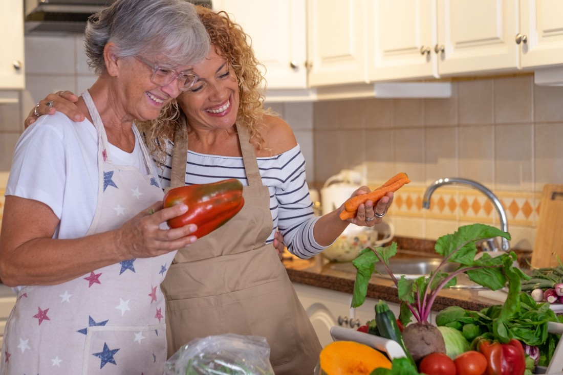 a woman and her elderly loved one stand in the kitchen with an array of healthy vegetables