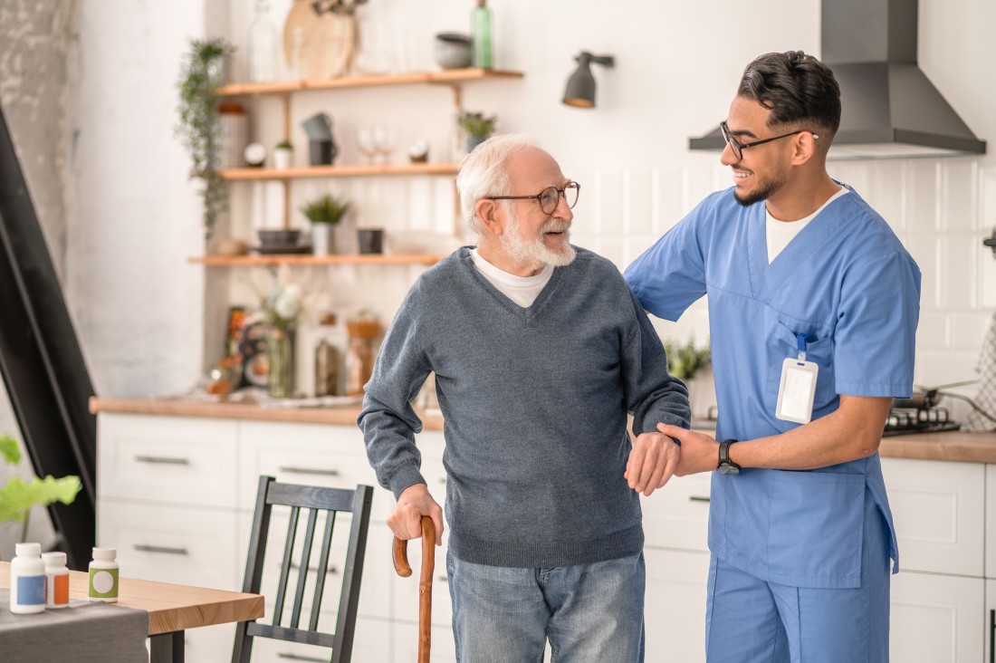 A caregiver helps an older man use a walking aid in his home kitchen